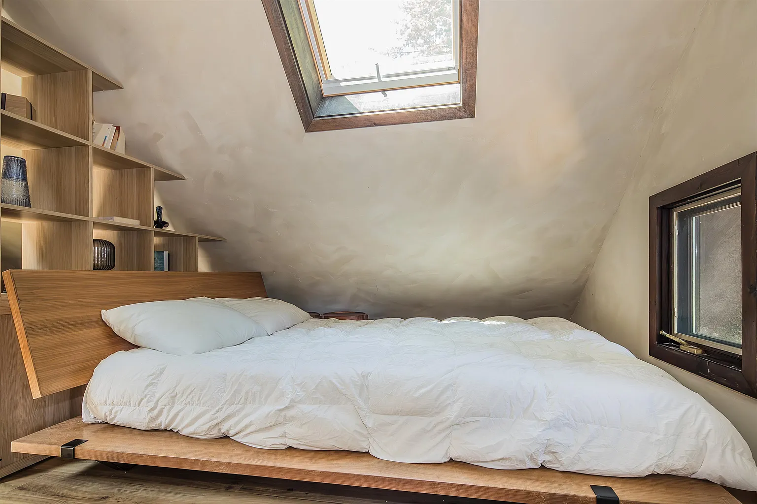The loft bedroom with a wooden bed, white bedding, built-in shelves, and a skylight